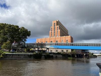 Looking+across+a+river+at+an+orange+brick+building+against+a+cloudy+sky.+A+blue+pedestrian+bridge+is+in+the+foreground.