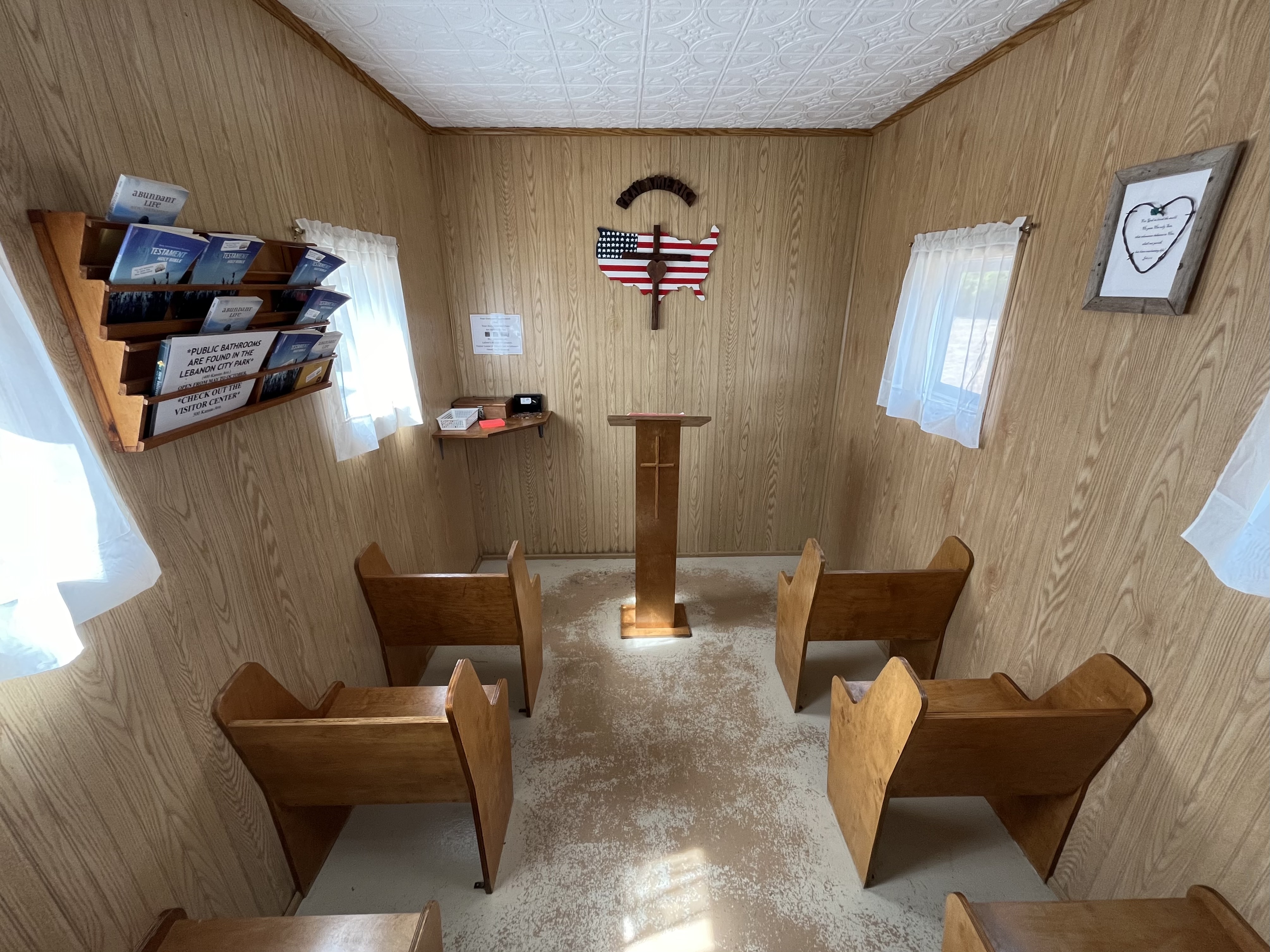 The+interior+of+a+small+chapel%2C+there+are+6+visible+seats%2C+a+small+lectern%2C+and+a+map+of+the+48+contiguous+states+painted+like+an+American+flag%2C+with+a+cross+and+a+heart+layered+over+it.+A+sign+saying+